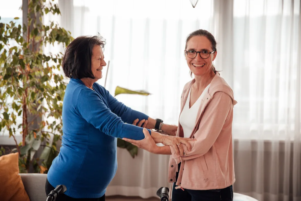 Die pflegende Angehörige hilft ihrer Mutter beim Aufstehen vom Sofa. Sie lächelt dabei in die Kamera.