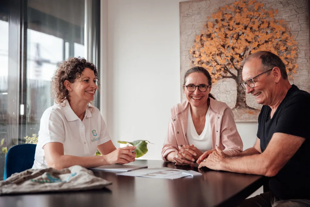 Eine Fachspezialistin von solicare klärt die pflegenden Angehörige und ihren Klienten beim persönlichen Gespräch am Tisch über die Vorteile einer Anstellung auf. Alle drei Personen sehen fröhlich aus.