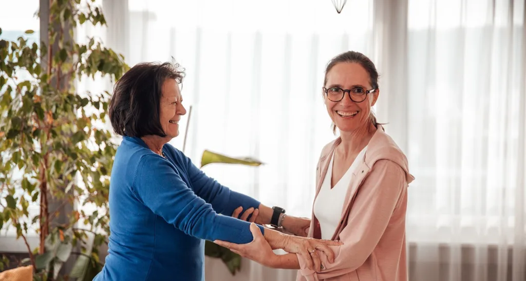 Die pflegende Angehörige hat Ihre Mutter beim Aufstehen unterstützt und hält sie fest an den Armen. Sie stehen sich vis-a-vis. Die pflegende Angehörige lächelt dabei in die Kamera.