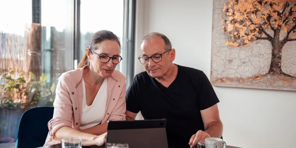 Die pflegende Angehörige sitzt zusammen mit ihrem Partner am Tisch und bedient ein Tablet. Auf dem Tablet ist der kostenlose Lehrgang QualiMio zu sehen.