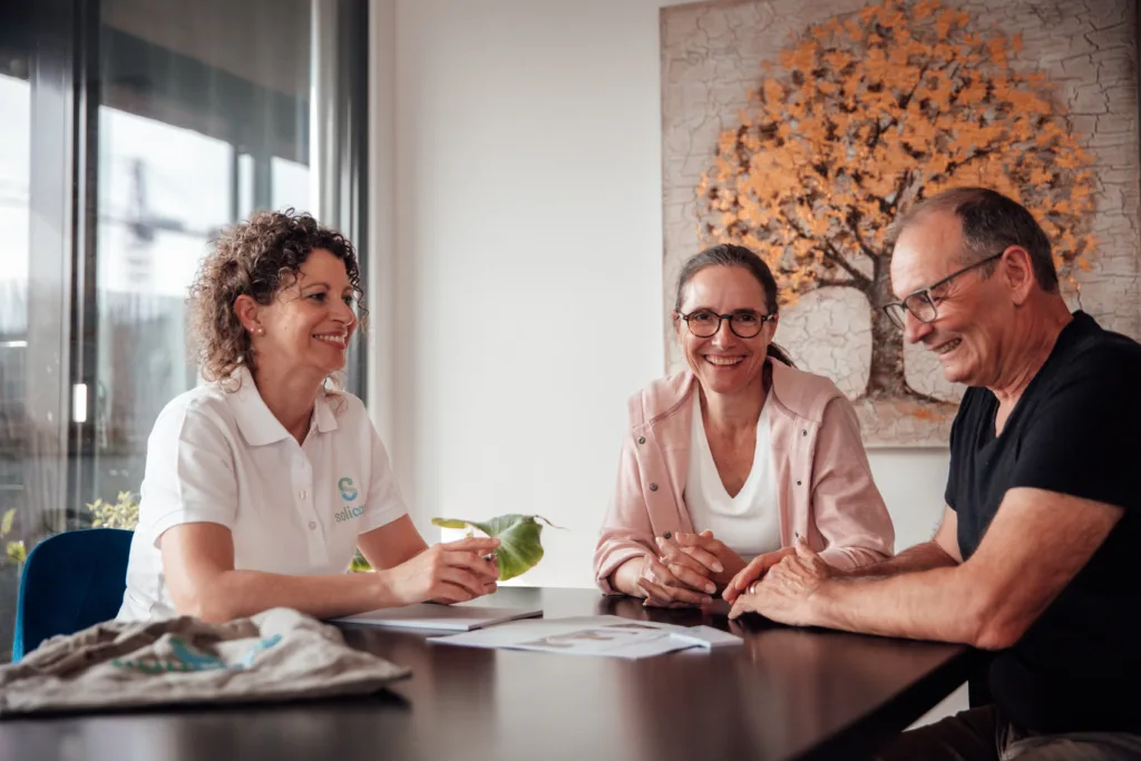 Eine Fallführende Pflegefachfrau von solicare sitzt am Tisch mit der pflegenden Angehörigen und dessen Klient. Sie klärt die beiden über die Vorteile einer Anstellung bei solicare auf.