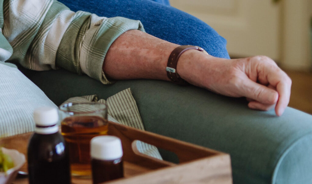 Eine Hand liegt auf der Sofalehne und auf einem Tablett steht ein Glas mit Tee und zwei kleinen braunen Apotheken-Fläschchen.